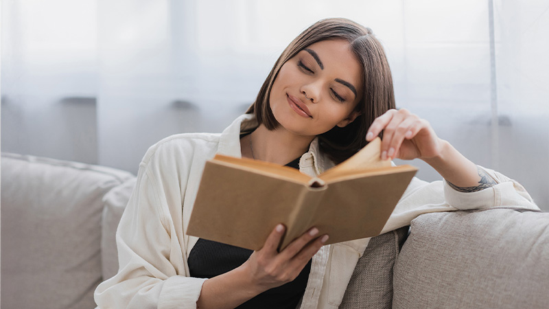 A person sits on a couch, reading a book and smiling, with light curtains in the background.