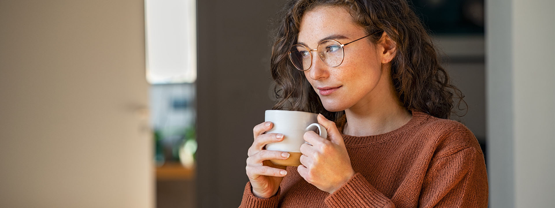 A person in a brown sweater holding a white mug close to their chest, standing indoors with a bright window and room interior in the background.