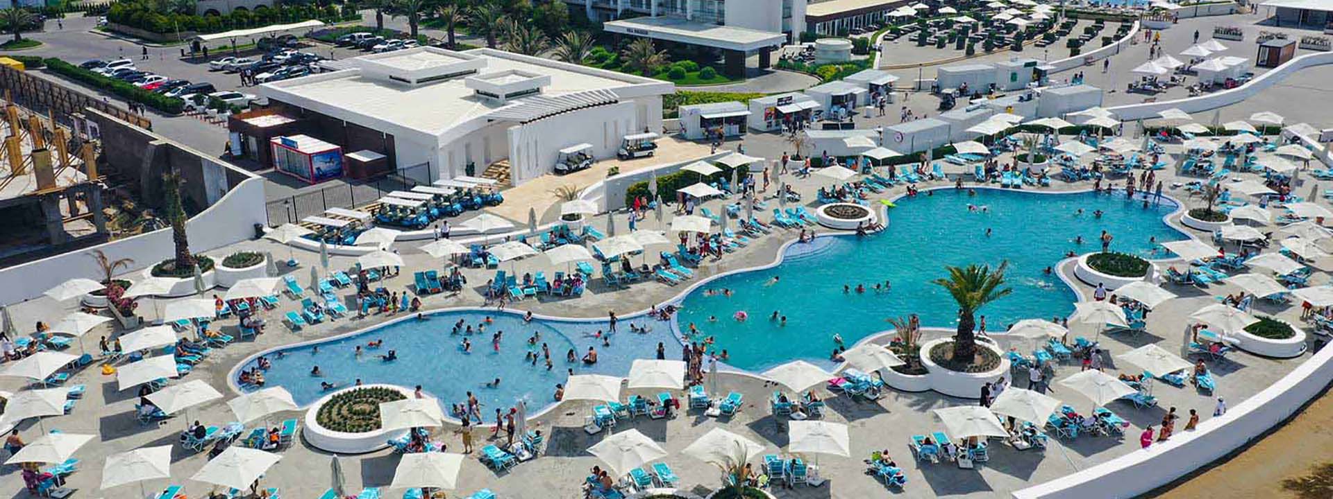 Large outdoor pool area with several sections of water, surrounded by lounge chairs and umbrellas, filled with people swimming and sunbathing. In the background, there are buildings and additional pool areas.