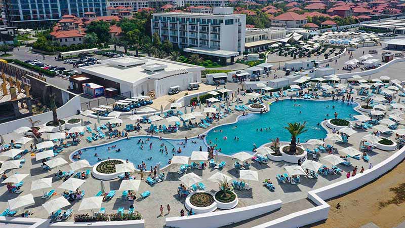 Large outdoor pool area with several sections of water, surrounded by lounge chairs and umbrellas, filled with people swimming and sunbathing. In the background, there are buildings and additional pool areas.