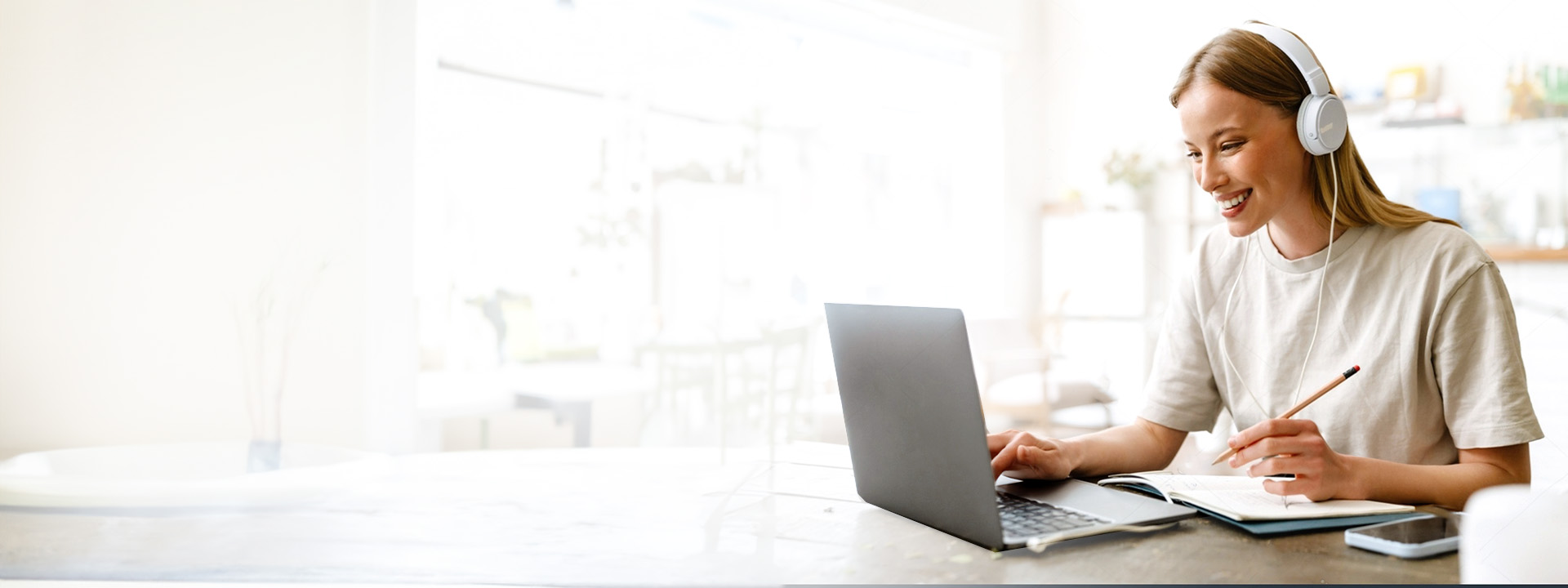 A young woman wearing headphones smiles while using a laptop and taking notes at a bright cafe table.