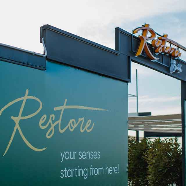Image of a sign with the word "Restore" in large cursive letters, accompanied by the phrase "your senses starting from here!" in smaller print. Another sign above reads “Restore” with decorative elements.
