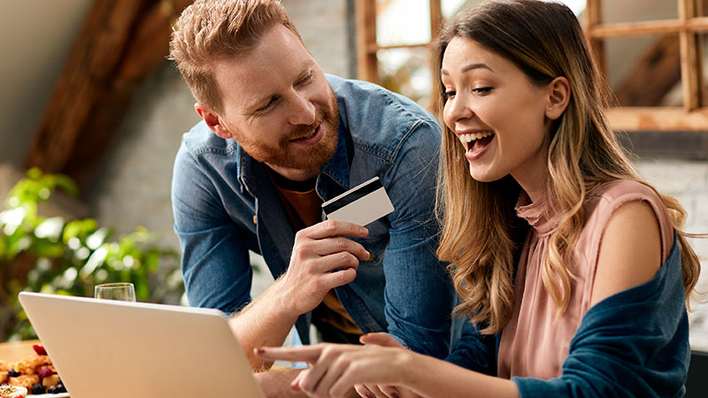 Two people are sitting together with a laptop. One person is holding a credit card and smiling while the other is pointing at the laptop screen.